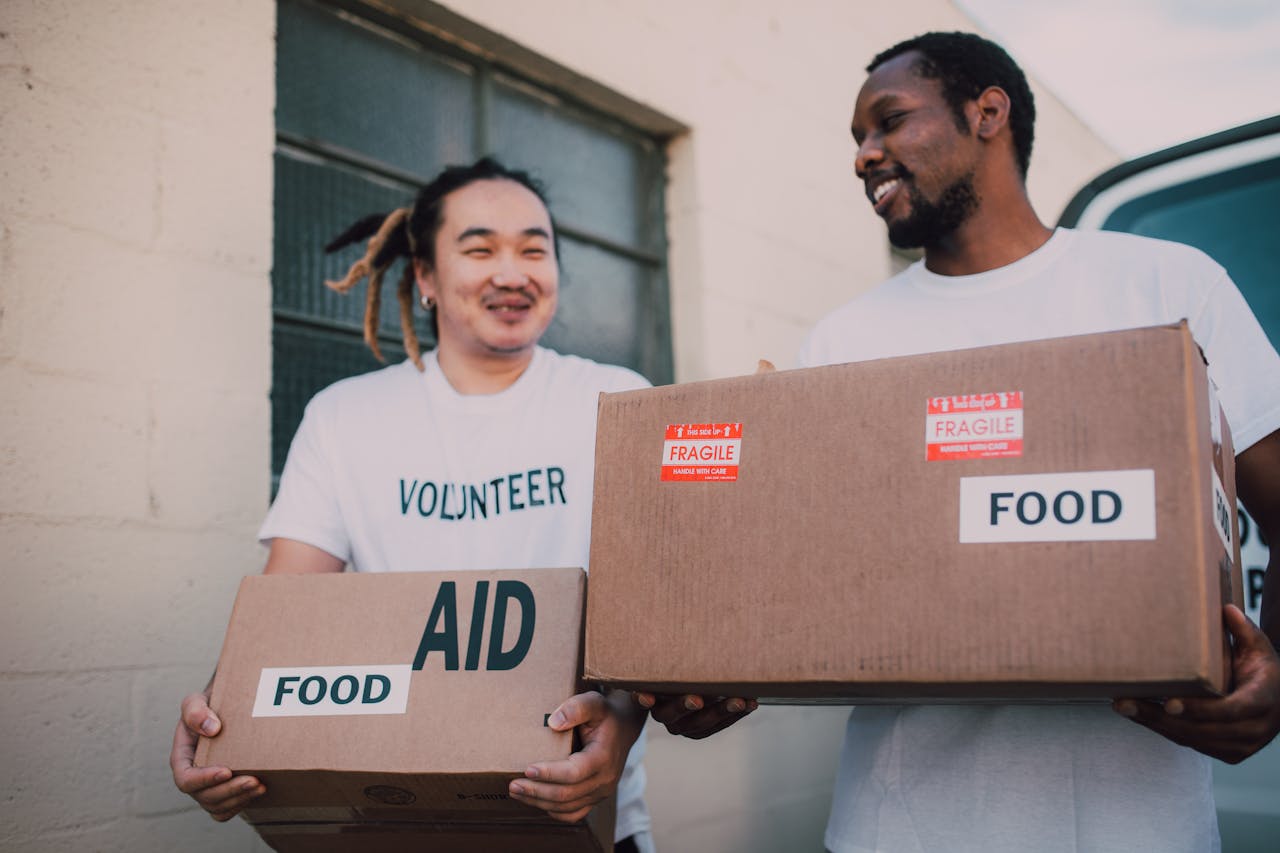 about-us Two volunteers carrying aid boxes labeled food, showing community support and charity efforts.